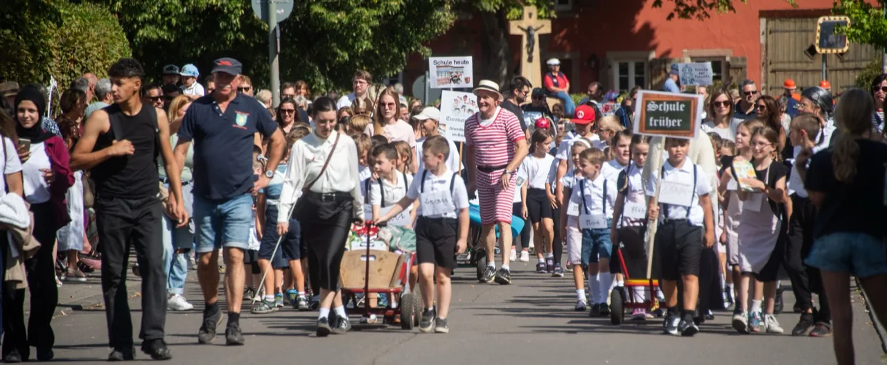 Eine große Gruppe von Schulkindern zieht mit historischen Schildern durch eine von Gästen gesäumte Straße.
